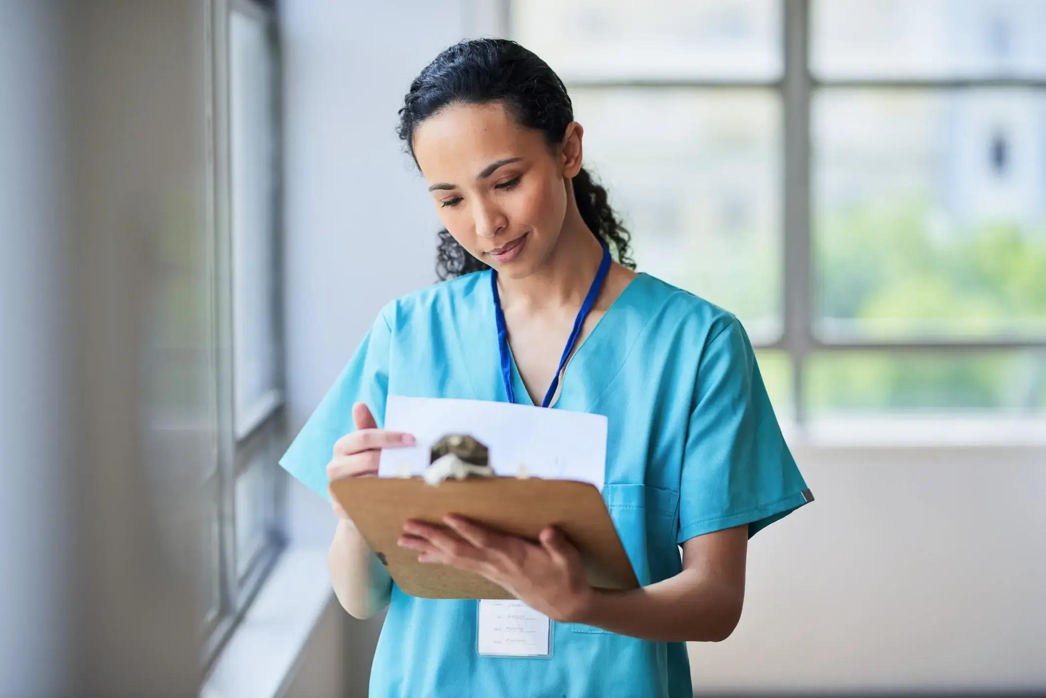 A healthcare professional with dark curly hair wearing blue scrubs and a lanyard, looking down at a wooden clipboard with papers in a bright office.